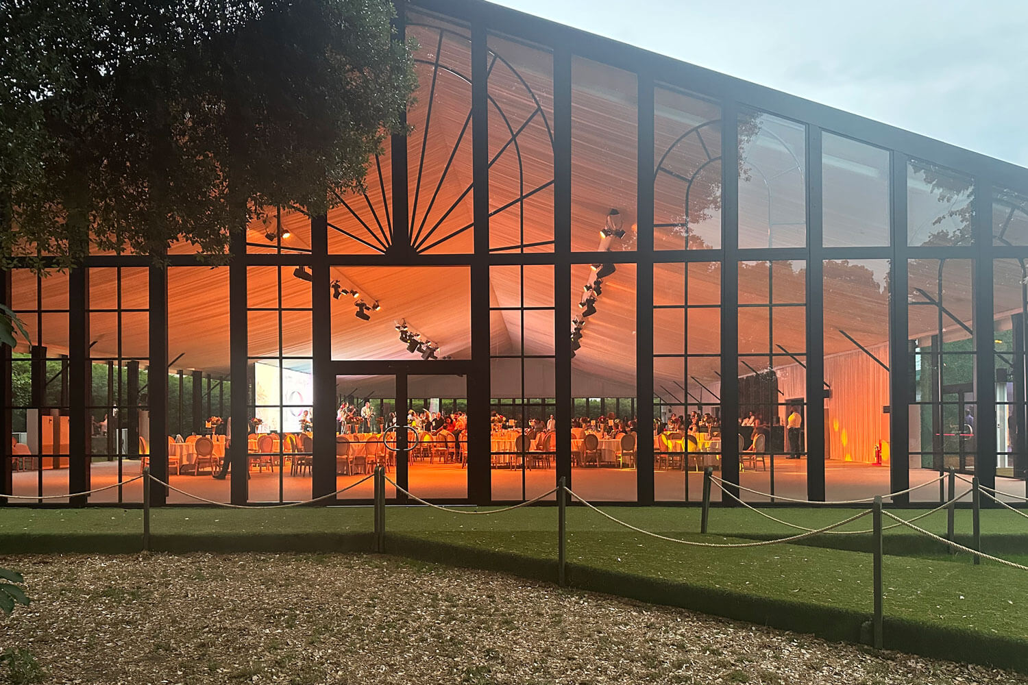 Large glass marquee at Syon Park with an event inside, reflecting trees and sky.
